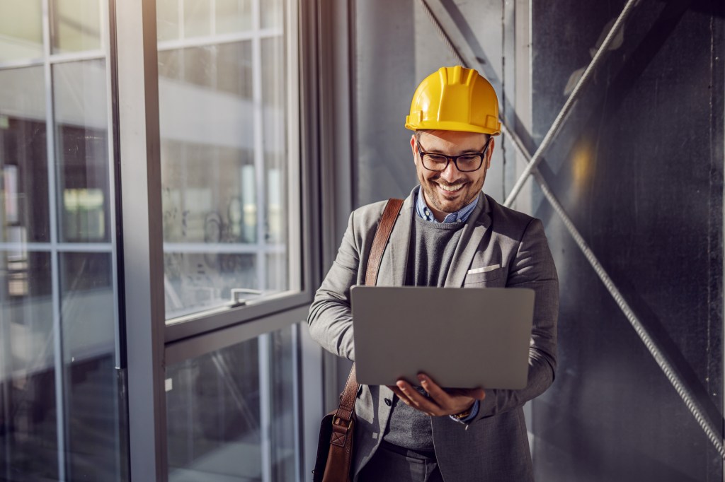 Young smiling architect standing near window in building in construction process and looking at blueprints on laptop.